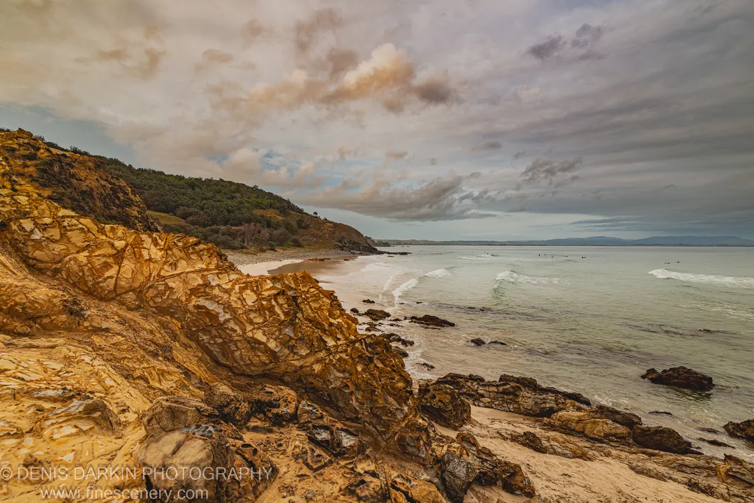 Stratified cliff rock in the foreground overlooks a curved sandy cove and calm ocean on the Queensland coast at dawn, under layered cloud.
