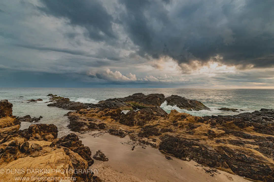 Ochre-toned coastal rock formations and algae-covered surfaces along the Queensland shoreline at dawn, with heavy storm cloud and a lit horizon break.