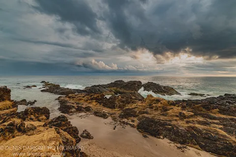 Ochre-toned coastal rock formations and algae-covered surfaces along the Queensland shoreline at dawn, with heavy storm cloud and a lit horizon break.
