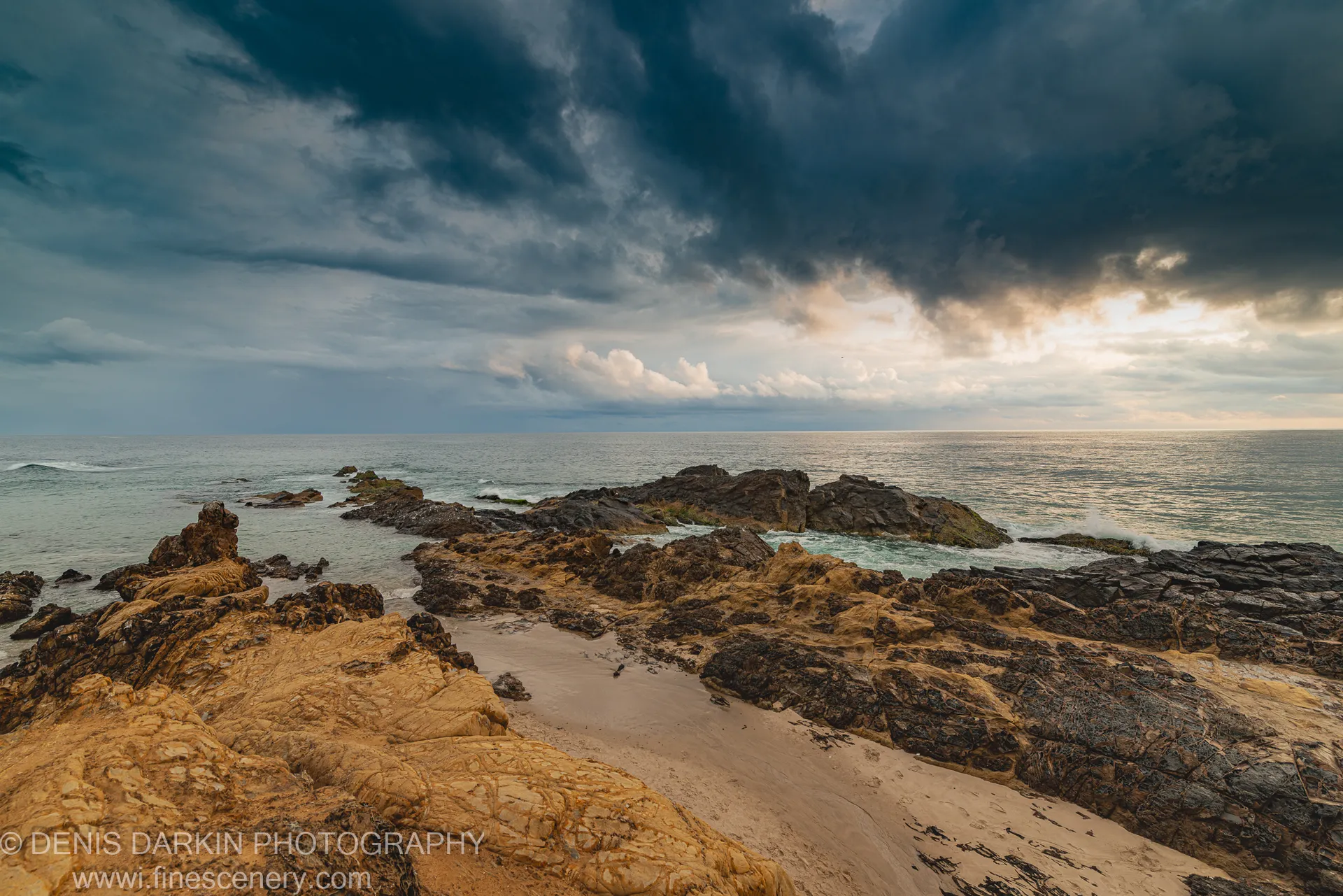 Amber-hued eroded sandstone platform on the Queensland coast at dawn, with dark storm clouds overhead and a warm break of light at the horizon.