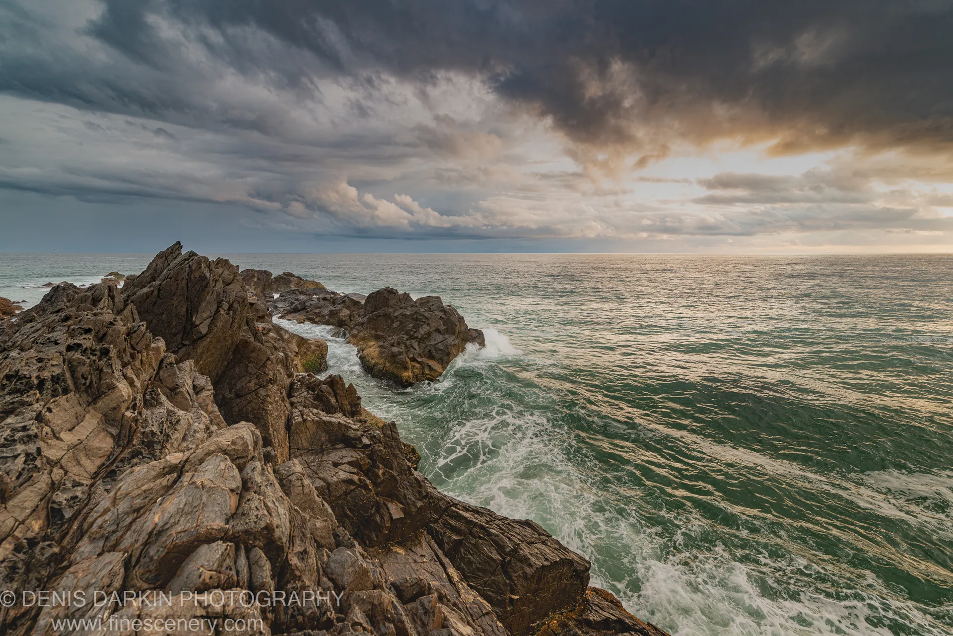 Weathered jagged rocks jutting into the Queensland ocean at dawn, with white-capped waves and a heavy storm bank overhead, warm light at the horizon.
