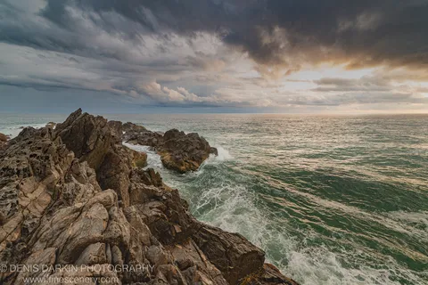 Weathered jagged rocks jutting into the Queensland ocean at dawn, with white-capped waves and a heavy storm bank overhead, warm light at the horizon.