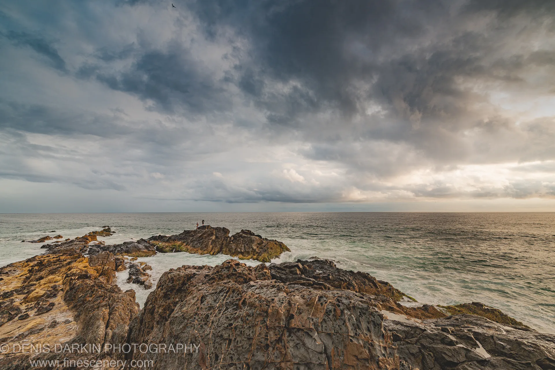Two silhouettes stand on a cliff edge above crashing surf on the Queensland coast, dark storm clouds overhead at dawn.