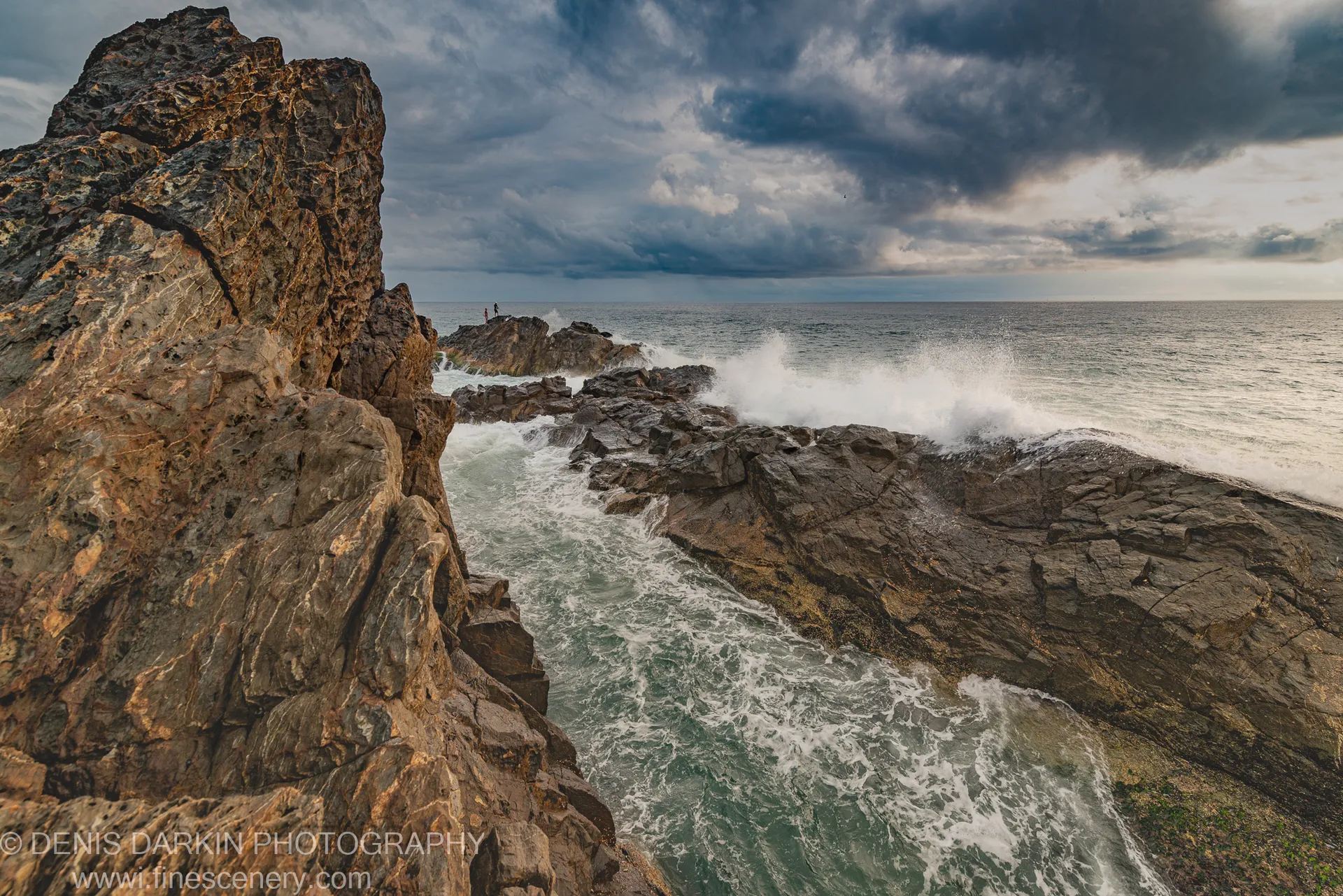 Surf crashes against sandstone columns on a Queensland headland at dawn, storm clouds above and two small figures visible on a distant jetty.