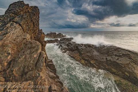 Surf crashes against sandstone columns on a Queensland headland at dawn, storm clouds above and two small figures visible on a distant jetty.