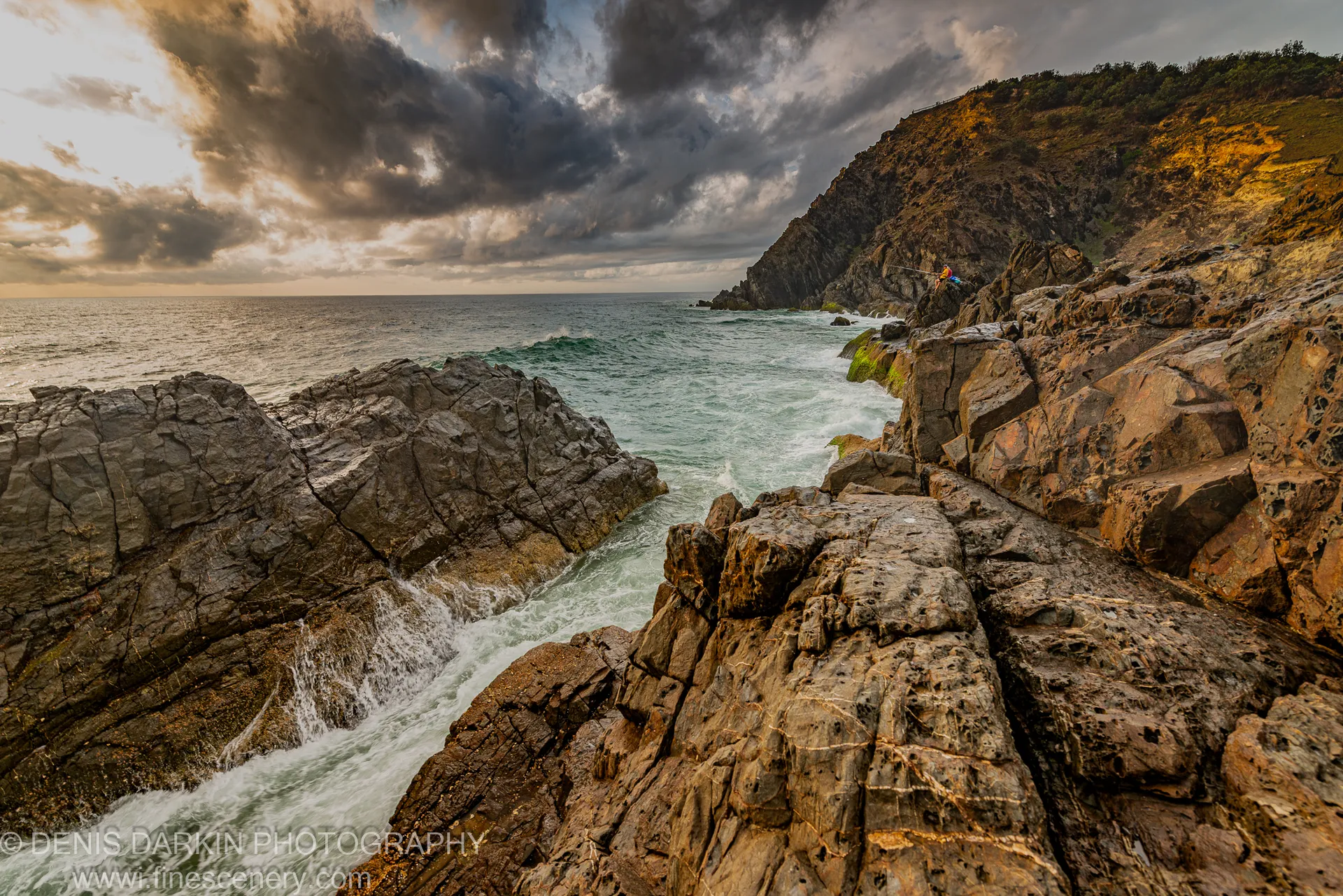 A lone figure stands high on a coastal path above fractured rock formations and crashing waves on the Queensland coast at dawn.