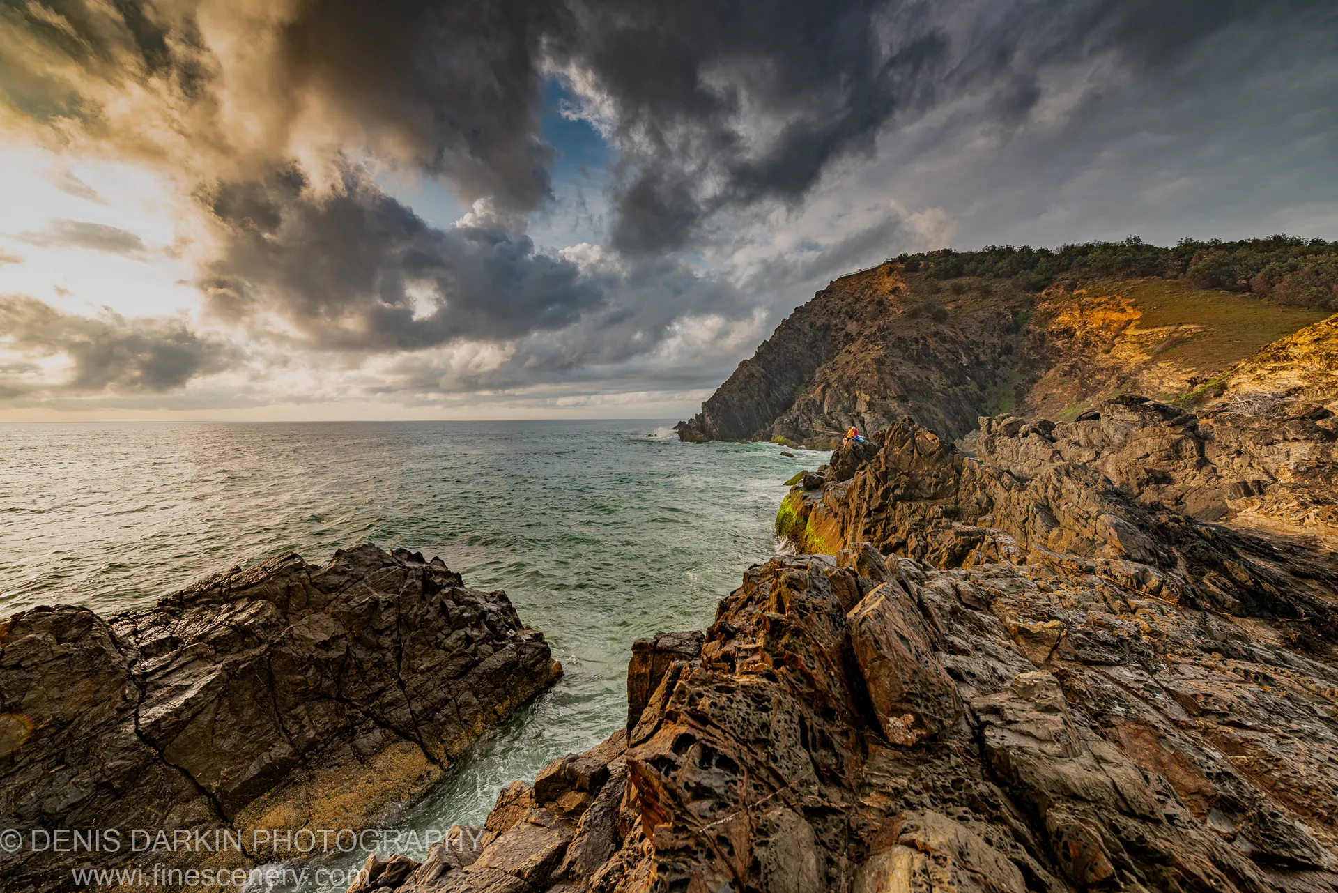 Weathered rocks in the foreground open onto a coastal bay in warm low-angle light beneath storm clouds, Queensland coast.