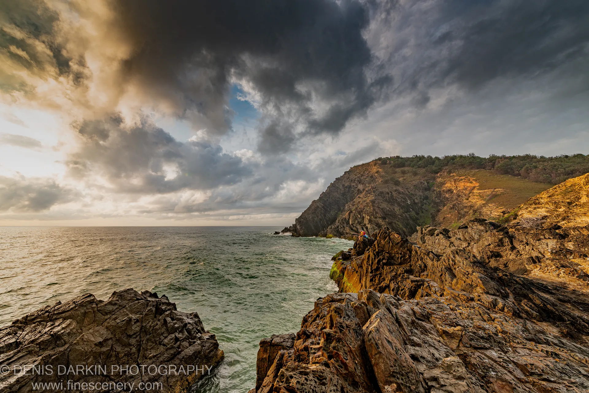 A solitary figure rests on a dark rocky headland at dawn on the Queensland coast, jagged stone in the foreground under heavy cloud.