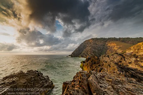 A solitary figure rests on a dark rocky headland at dawn on the Queensland coast, jagged stone in the foreground under heavy cloud.