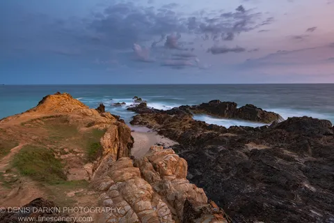 Volcanic rock formations at a sandy cove, Byron Bay headland, with a long-exposure ocean and a predawn sky shifting from deep blue to pink.
