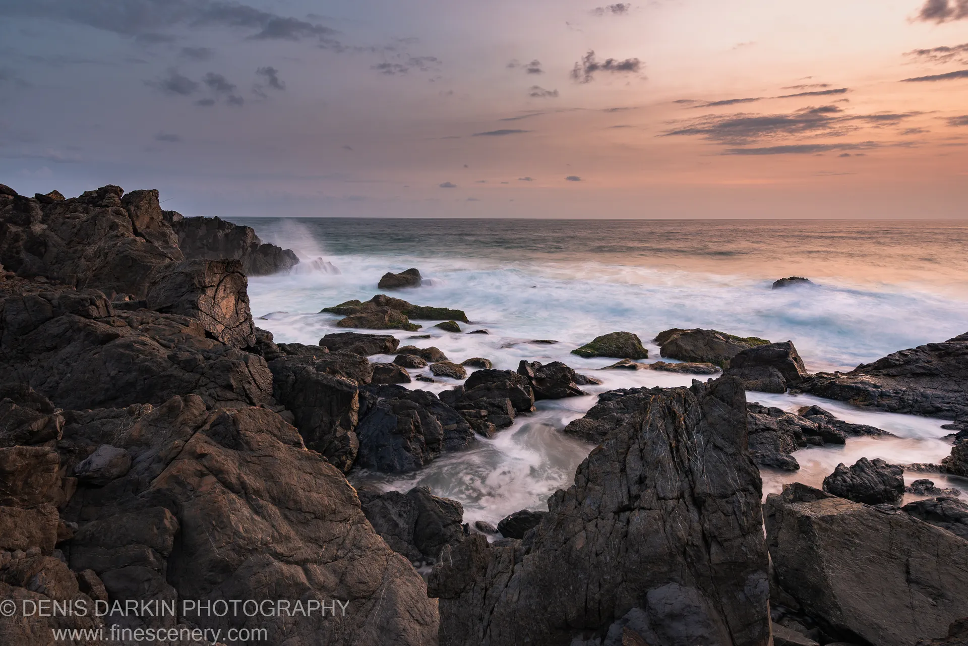 Dark moss-covered rocks anchor the foreground as long-exposure surf rolls across a rocky Queensland shore in the minutes before sunrise.
