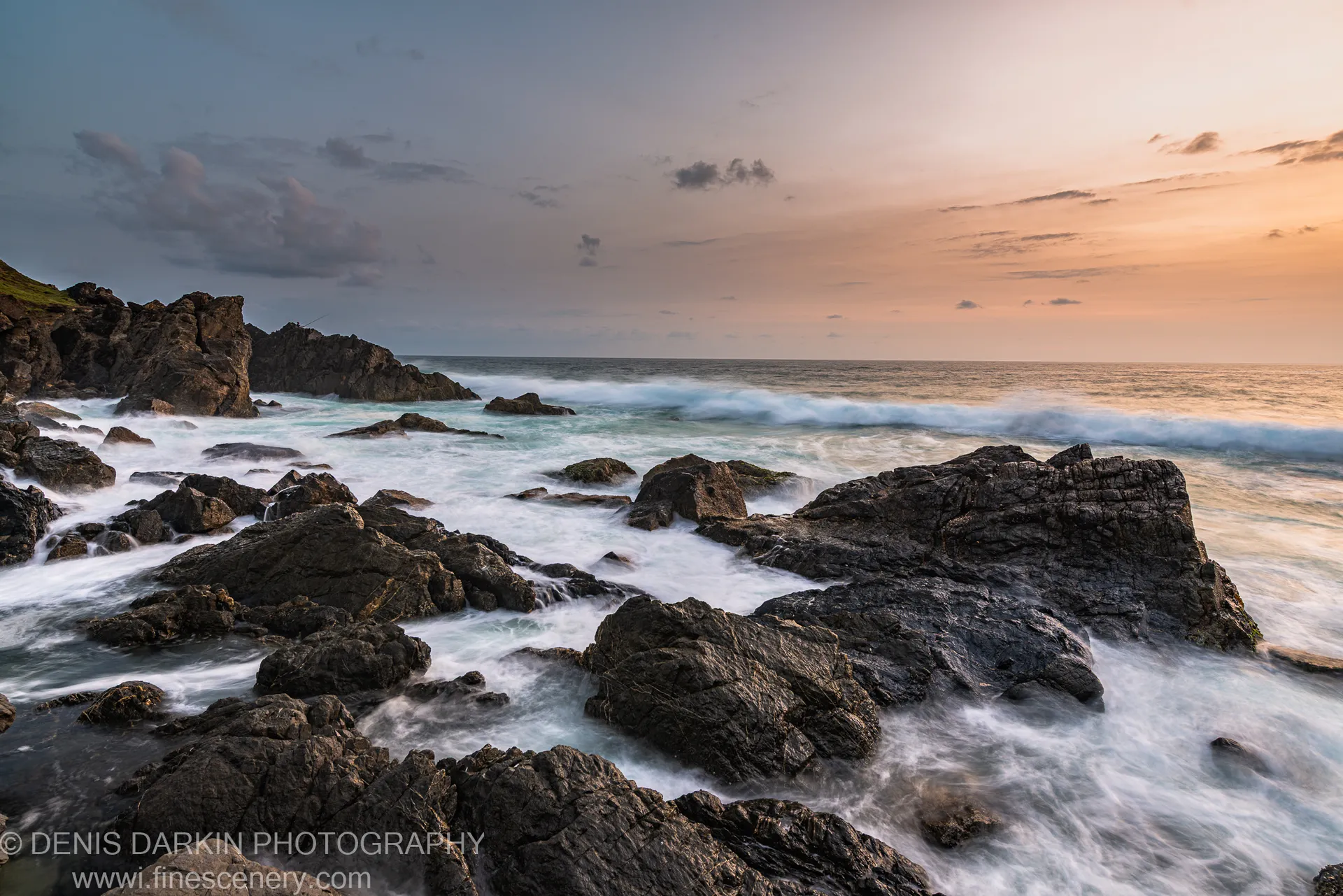 Wet textured rock formations on the Queensland coast reflect the gradient sunrise sky, scattered clouds and a grassy slope visible at the left edge.