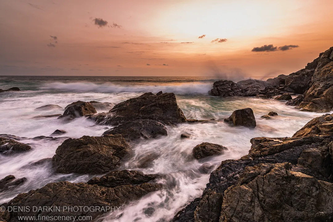 Long-exposure waves spreading as white foam across dark basalt boulders at the Byron Bay headland, with a warm glow on the predawn horizon.