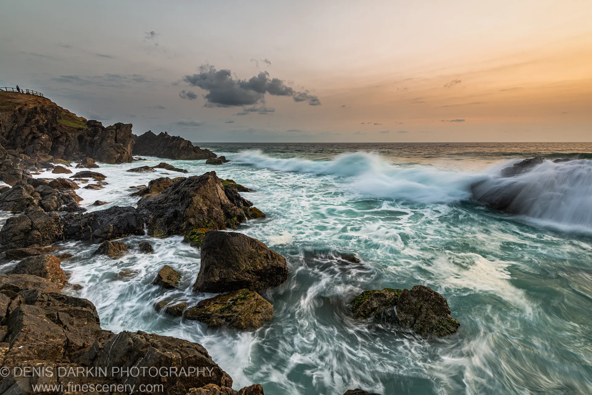 White foam swirling around mossy sea boulders at the Byron Bay headland, with a warm golden light on the right horizon before sunrise.