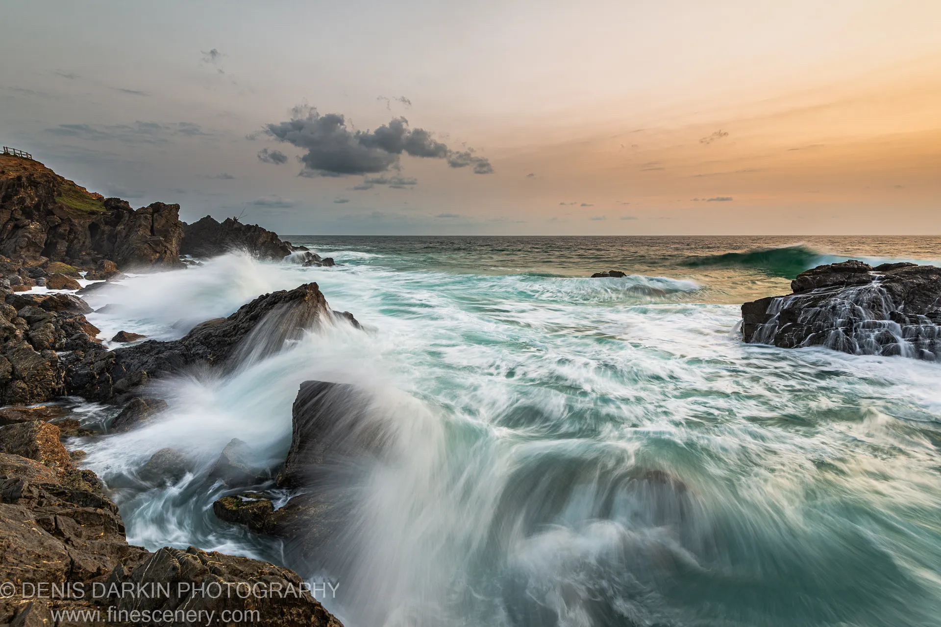 Dark coastal rocks emerge through a rush of long-exposure tidal water on the Queensland coast, beneath a sky warming from gold to grey.