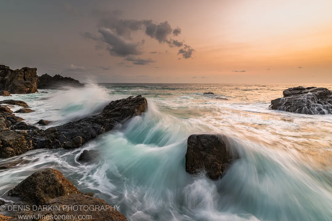 A breaking wave rolling over a basalt shelf at the Byron Bay headland before dawn, with warm golden light on the right horizon.