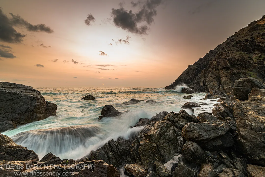 Rough coastal rocks and blurred surf in the foreground below a headland cliff, with an apricot sky at dawn, Queensland coast.