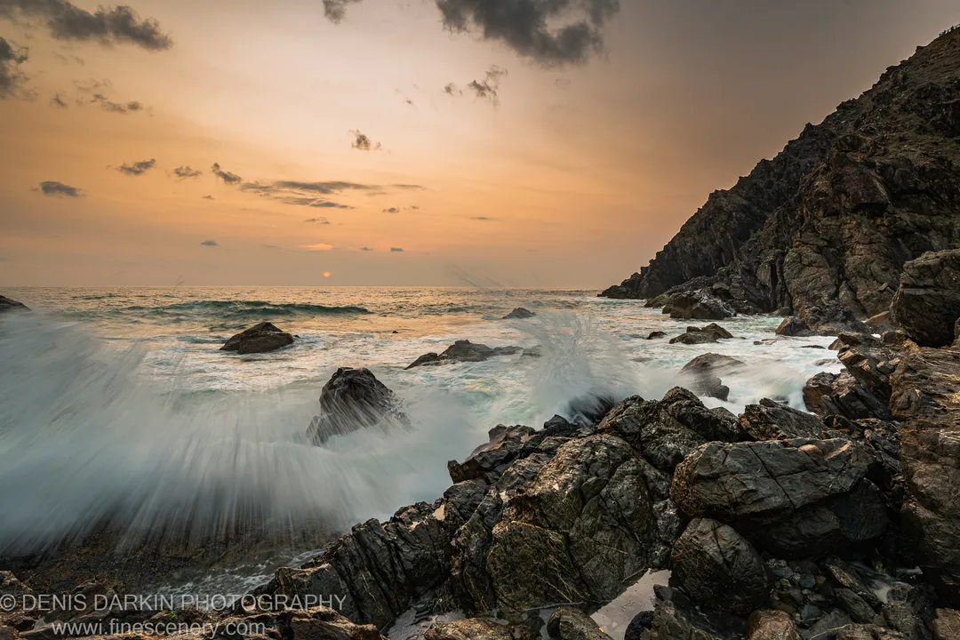 Jagged dark rocks on the Queensland coast at dawn, crashing waves rendered as white foam by long exposure, a cliff face rising on the right.
