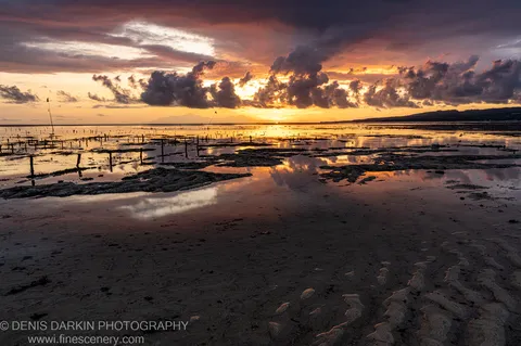 Wooden posts rise from tidal mudflats at low tide on the Bali coast, with heavy cloud formations and a mountain silhouette on the horizon.