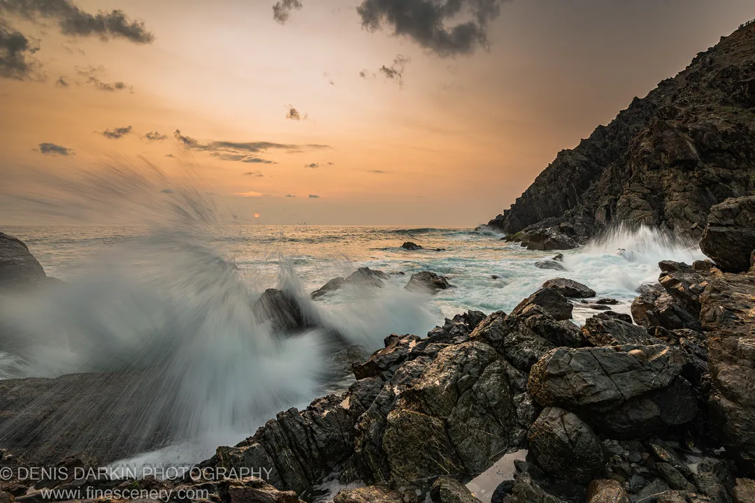 A tall cliff face on the right side of the frame at the Byron Bay headland at dawn, with long-exposure spray across the foreground rocks.