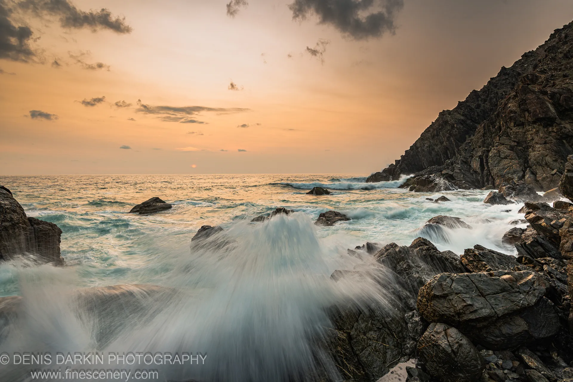 Dark coastal rocks and crashing waves at the base of a massive cliff, Queensland coast, with an orange-to-dark dawn sky above.