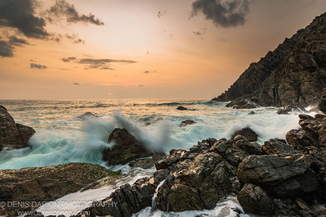 Mossy dark rocks on a Queensland shoreline at dawn, slow-shutter surf around the foreground stones and a steep cliff on the right.