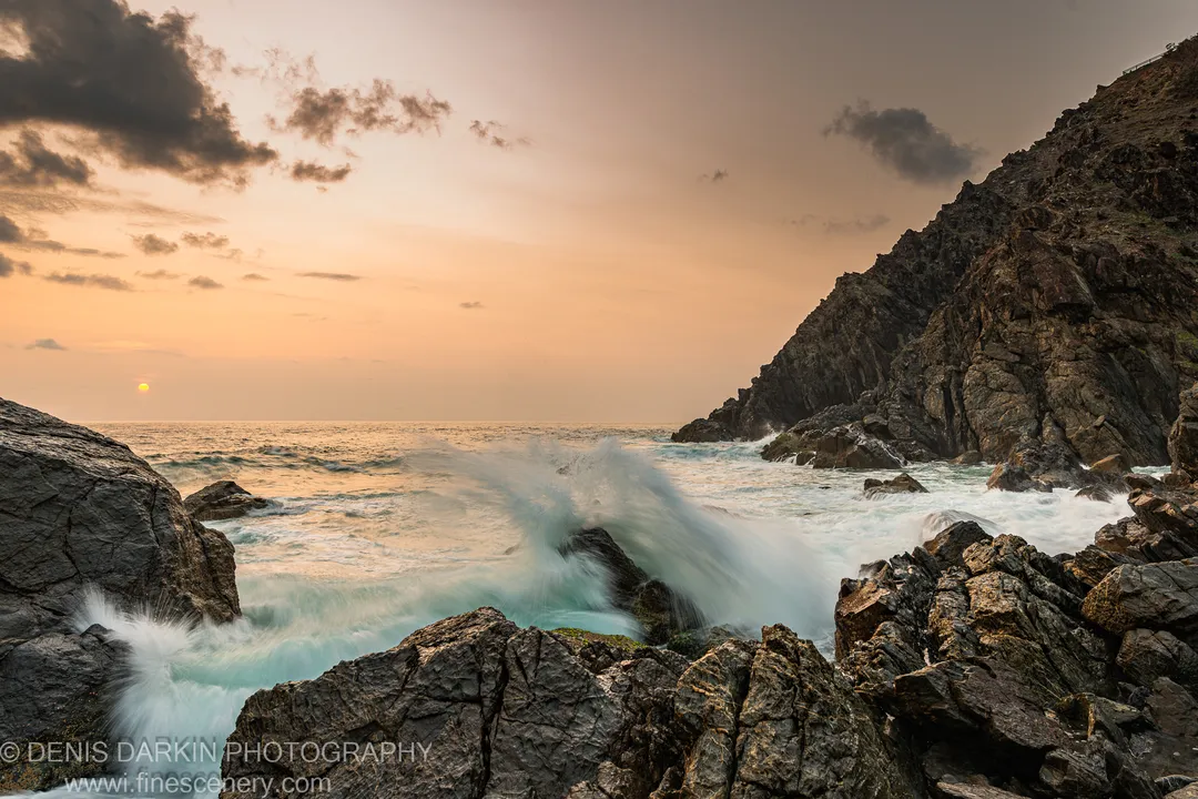 Turbulent surf crashing against jagged dark rocks on the Queensland coast at dawn, the sun as a small orb through mist on the horizon.