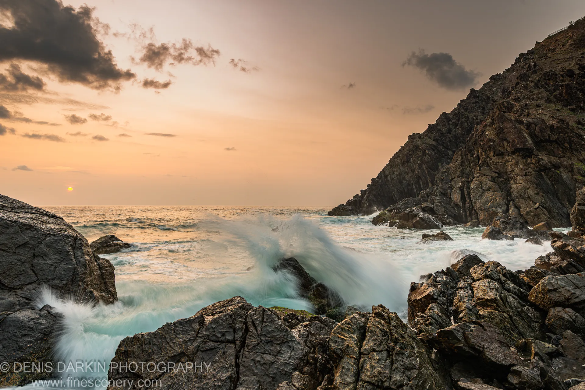 Turbulent surf crashing against jagged dark rocks on the Queensland coast at dawn, the sun as a small orb through mist on the horizon.