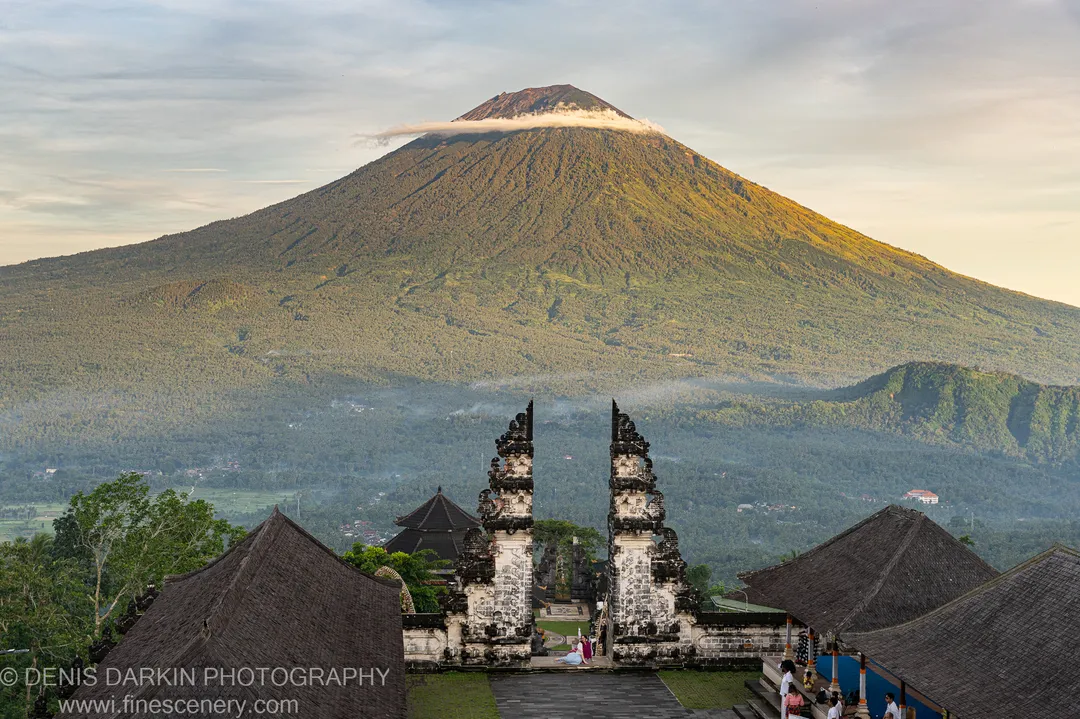 Stone gateways and dark temple rooftops stand at the base of a forested Balinese volcano whose summit is wrapped in cloud.