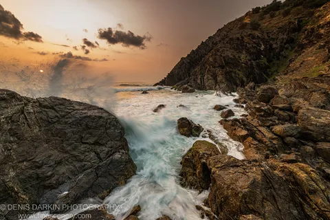 A column of sea spray rising against the dark cliff face at Byron Bay, with long-exposure water flowing smoothly through the basalt rocks.