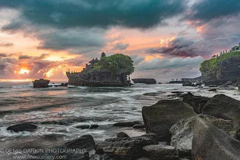 A tiered temple on a rocky ocean islet beneath storm clouds and a band of warm sunset light, Bali coast.