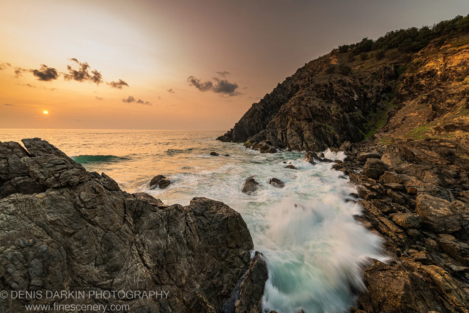 Rugged coastal cliff lit by early dawn light on the Queensland coast, with waves breaking against dark foreground rocks and scattered clouds above.