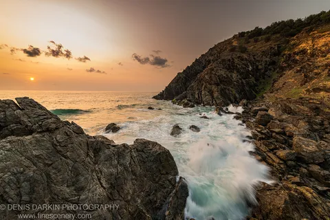Rugged coastal cliff lit by early dawn light on the Queensland coast, with waves breaking against dark foreground rocks and scattered clouds above.