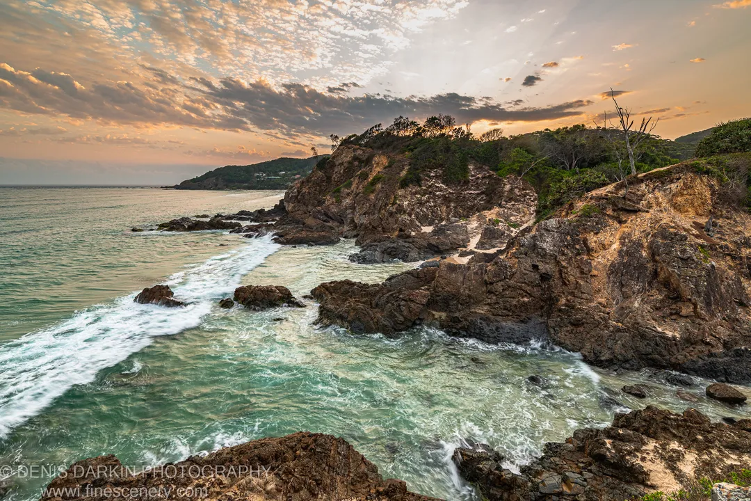 Sunlight breaking through cloud layers above the Byron Bay headland at dawn, with turquoise water foaming against dark rocks along the curved shoreline.