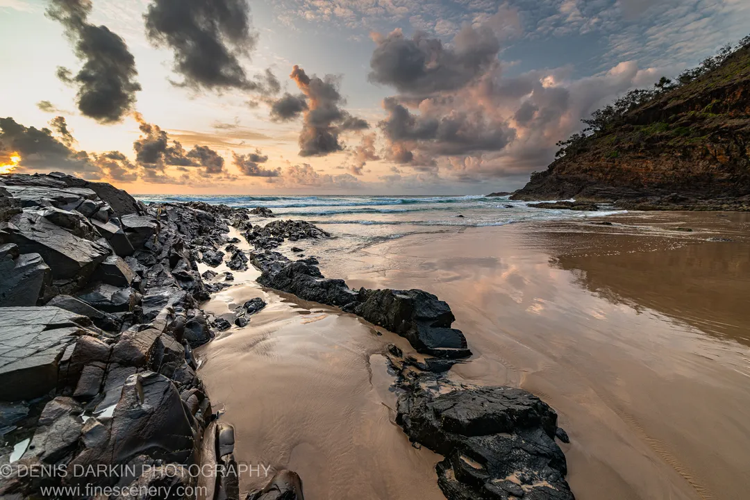 Dark angular rocks in the foreground of a coastal bay at Byron Bay, with wet sand reflecting a clouded dawn sky mixing grey and pink.