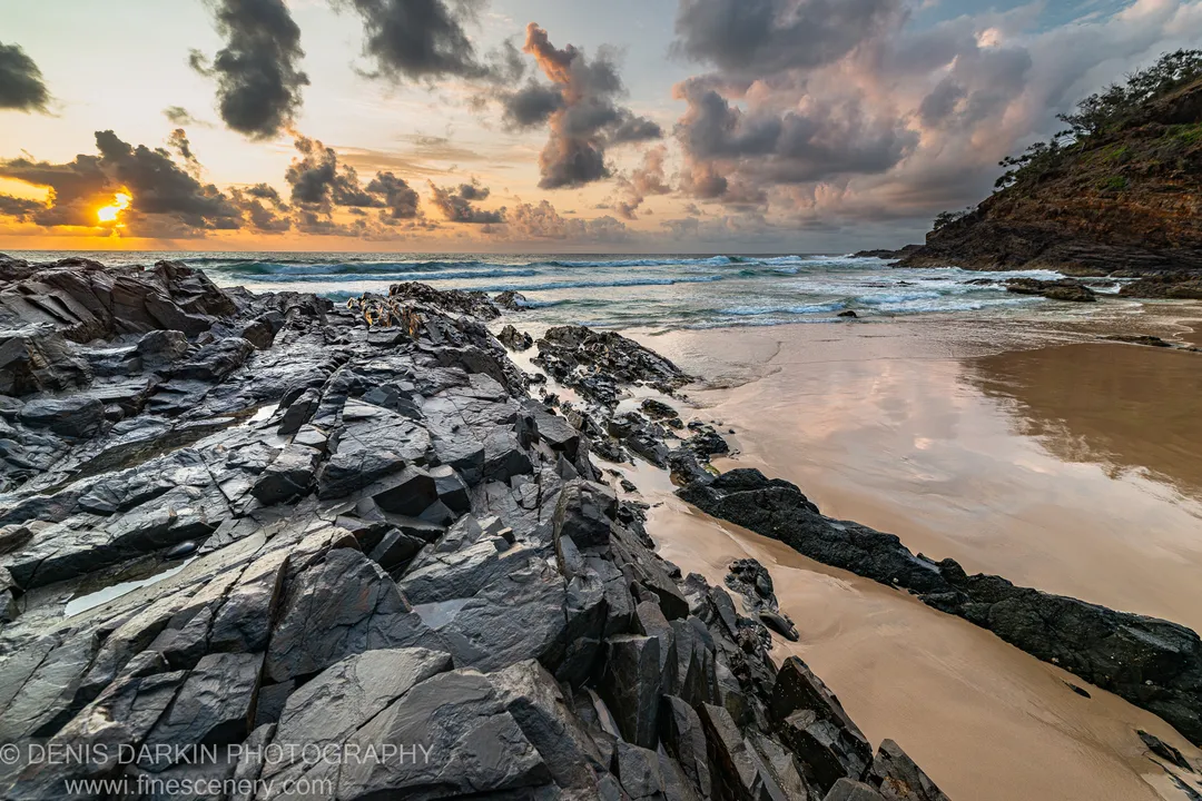 Fractured dark rock layers and wet sand reflect dawn light below scattered clouds at Byron Bay, Queensland.