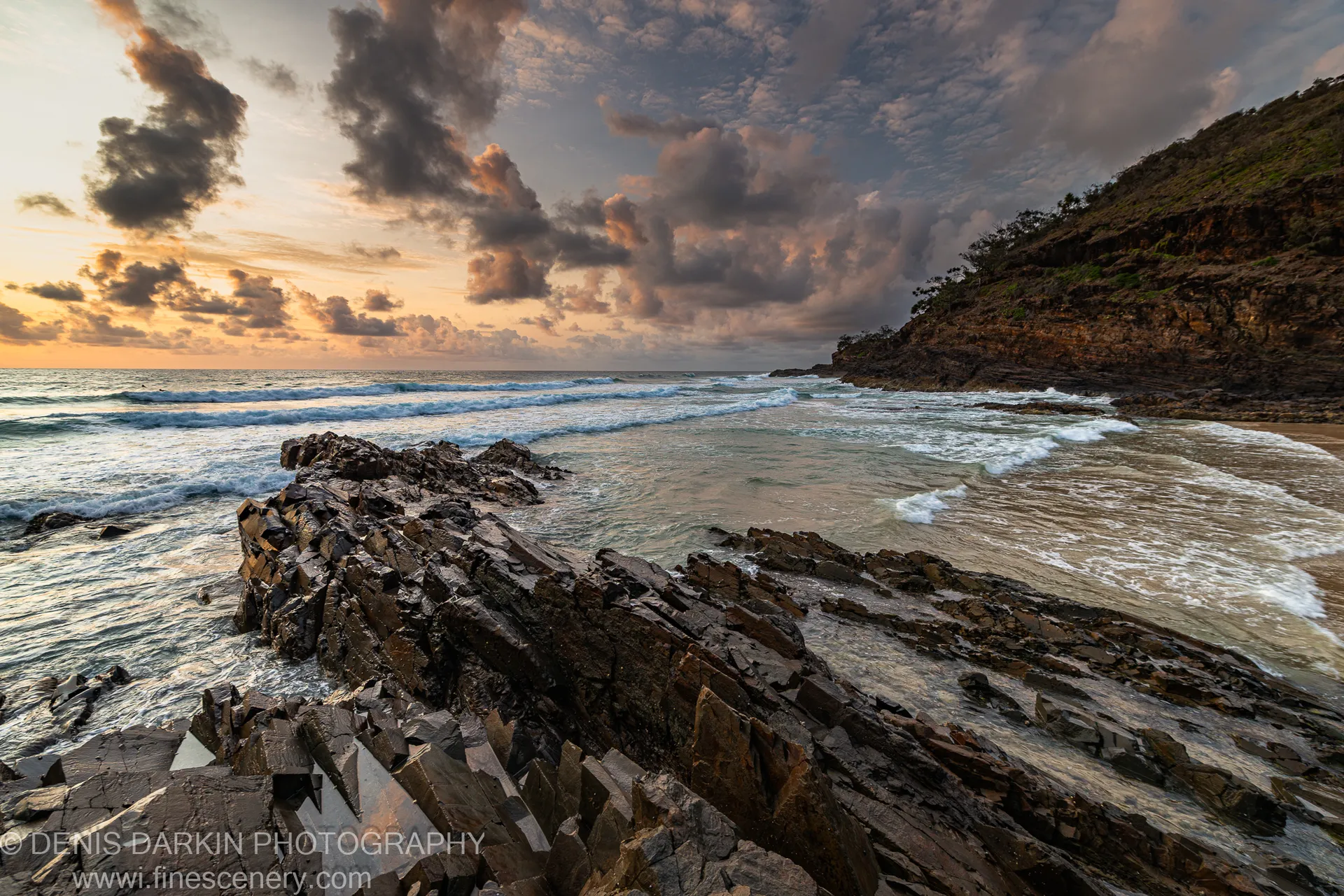 Jagged coastal rocks anchor the foreground as sunrise light breaks through heavy clouds above the Byron Bay headland.