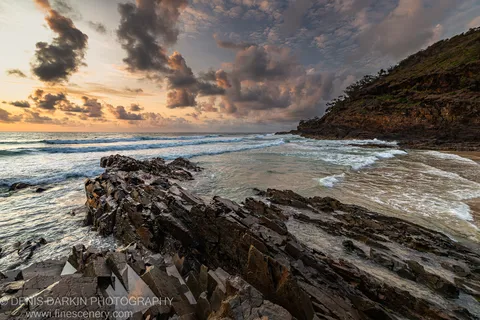 Jagged coastal rocks anchor the foreground as sunrise light breaks through heavy clouds above the Byron Bay headland.