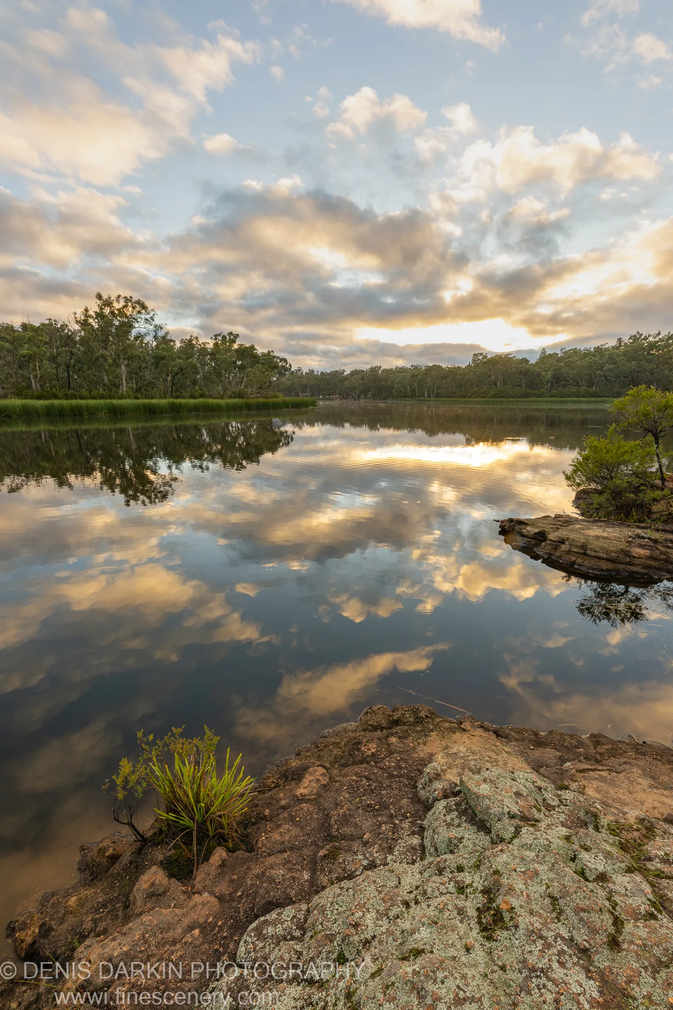 Lichen-covered rocks in the foreground of Dunns Swamp, New South Wales, with calm water reflecting textured clouds at low-angle light.