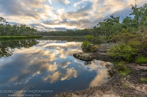 Still water at Dunns Swamp reflects warm and grey clouds, bordered by textured rocks and green forest.