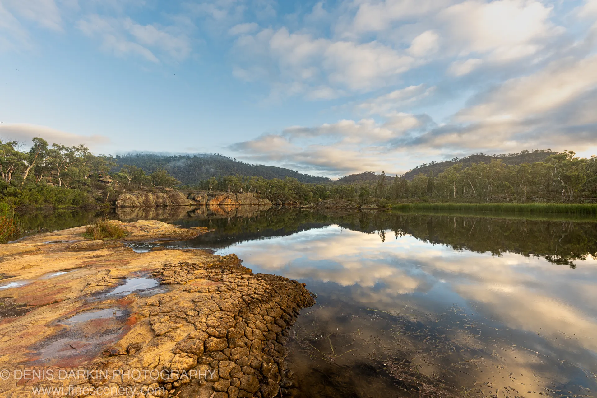 Polygonally cracked rocks in the foreground at Dunns Swamp, New South Wales, with calm water reflecting clouds and forested hills beyond.