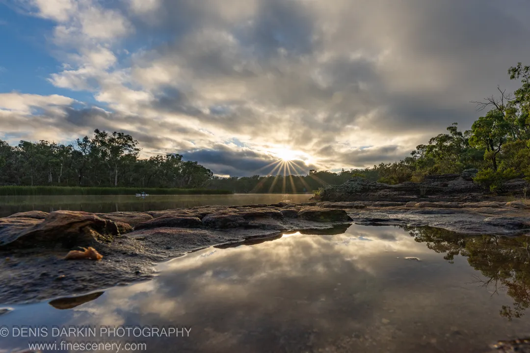 Sunburst through heavy clouds reflected in a shallow pool at Dunns Swamp, with green trees lining both banks.
