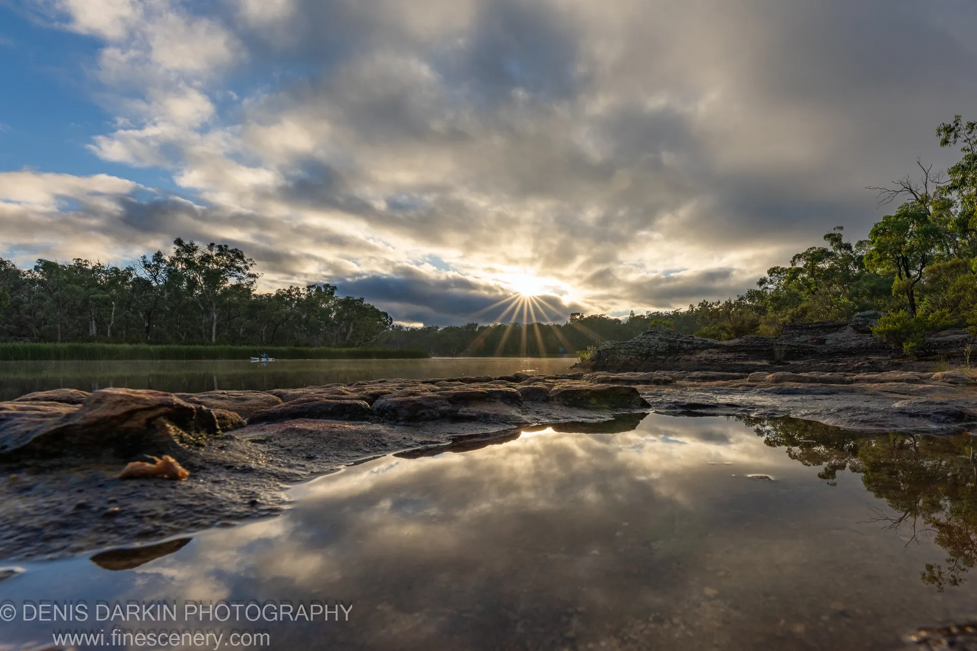 Sunburst through heavy clouds reflected in a shallow pool at Dunns Swamp, with green trees lining both banks.