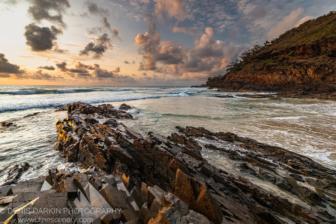 Fractured rock formations with layered textures on the Queensland coast at dawn, with a sheltered cove and vegetation-covered cliff visible beyond the breaking surf.