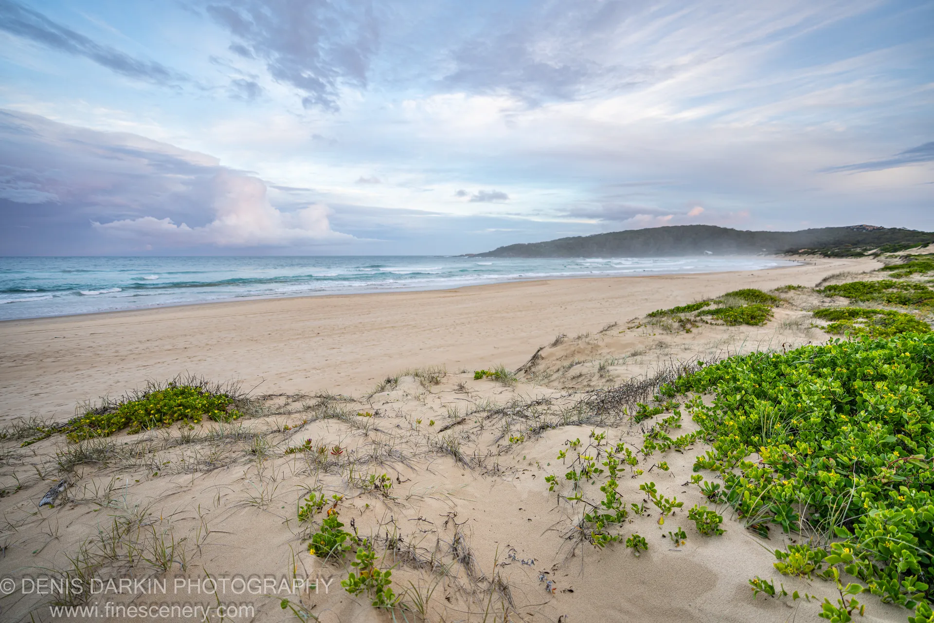 A tree-lined headland curves away beyond sand dunes and gentle white-capped rollers under a streaked pink afternoon sky at One Mile Beach.