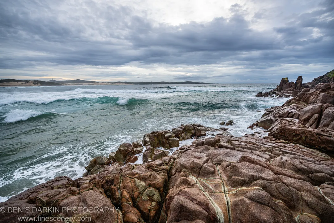 Massive reddish-brown rock formations anchor the foreground of a dawn seascape at One Mile Beach, Port Stephens, with a sandy headland stretching across the distance.