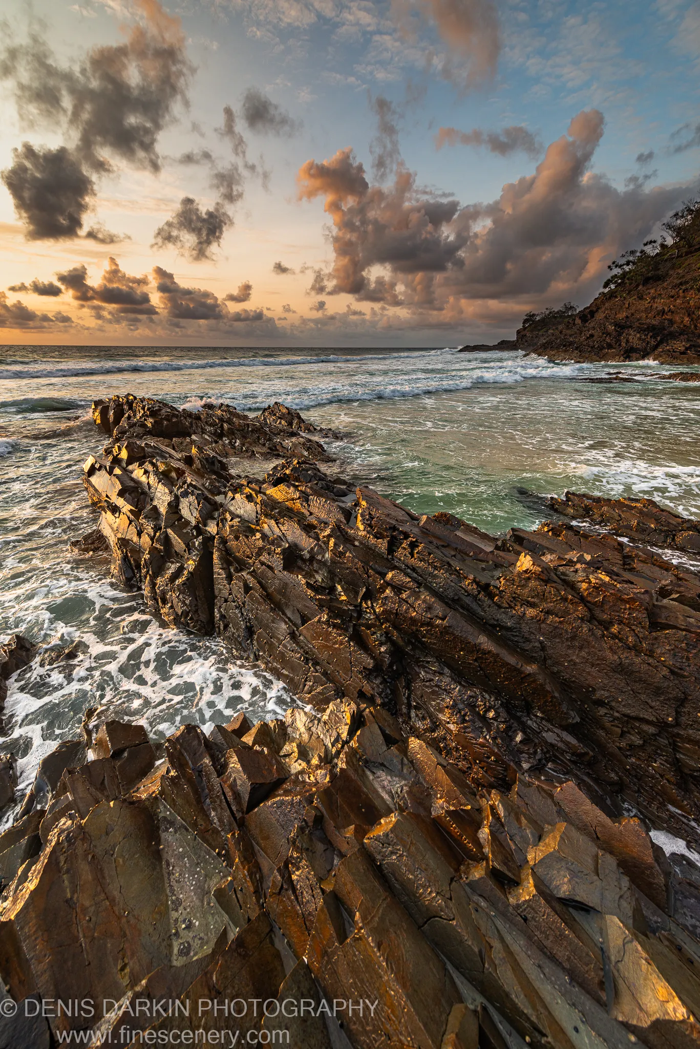Tilted rock strata run diagonally from the foreground to the surf at dawn, with sea-foam and massed cloud above, Byron Bay.