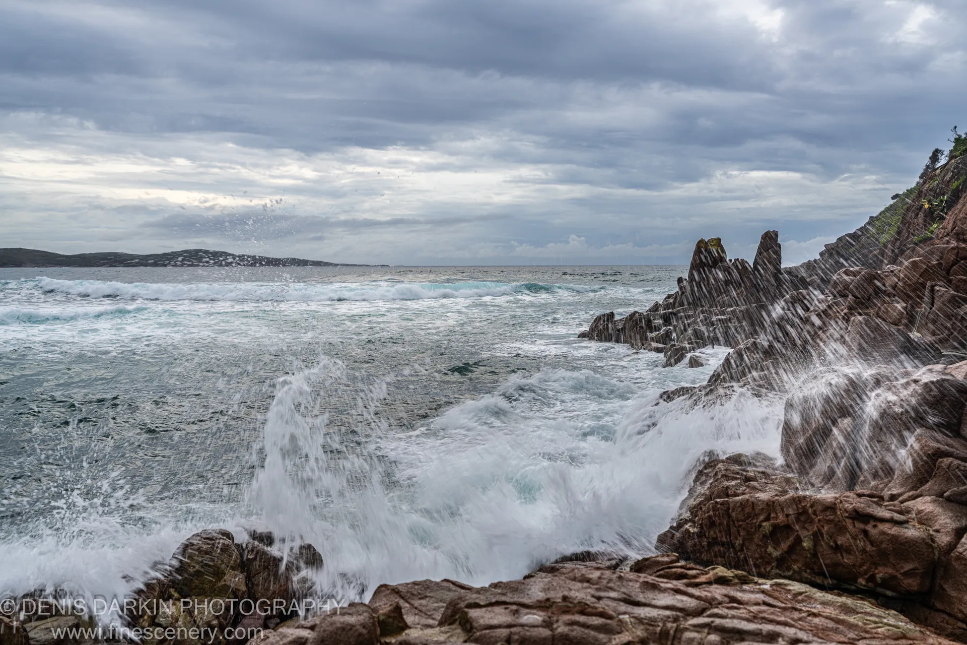 Turbulent surf crashes against rugged reddish-brown cliff rocks at dawn on One Mile Beach, sending white sea spray into the overcast air.