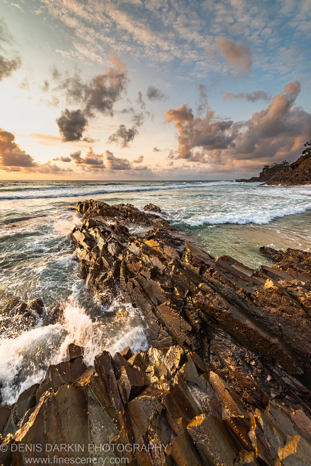 White sea-foam from a breaking wave contrasts against dark stratified rock and calm green water in a sheltered cove.