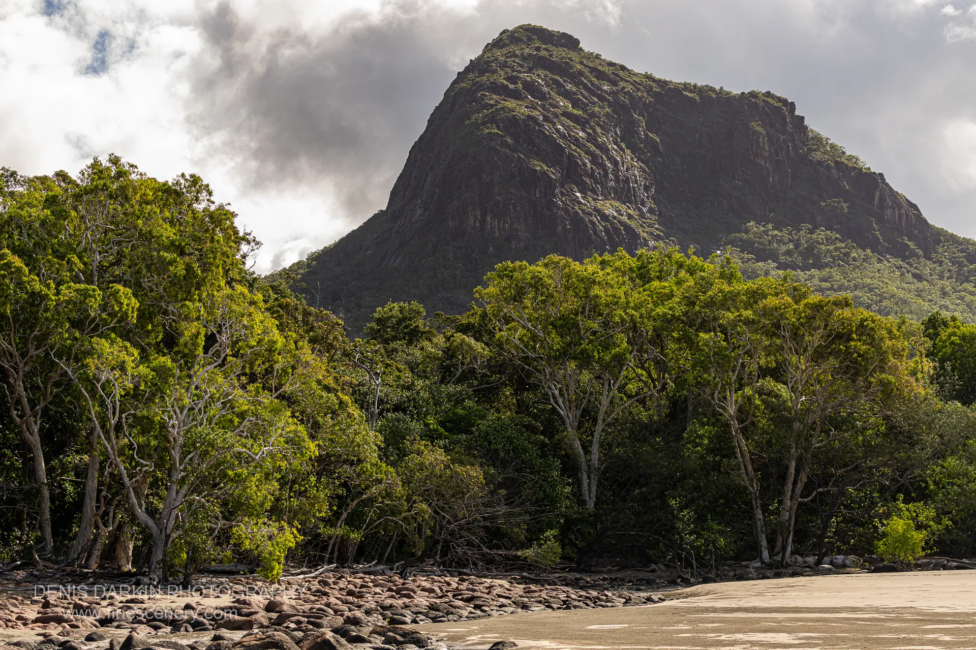 Dark granite dome of Nina Peak rises behind pale-trunked paperbark trees, with a boulder-strewn creek mouth running across the foreground sand.